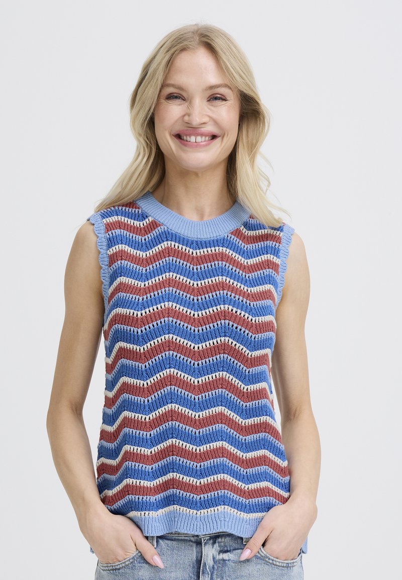 Femme souriante portant un haut sans manches en tricot avec des rayures zigzag horizontales bleu, marron et blanc, mains dans les poches de jean, fond clair.