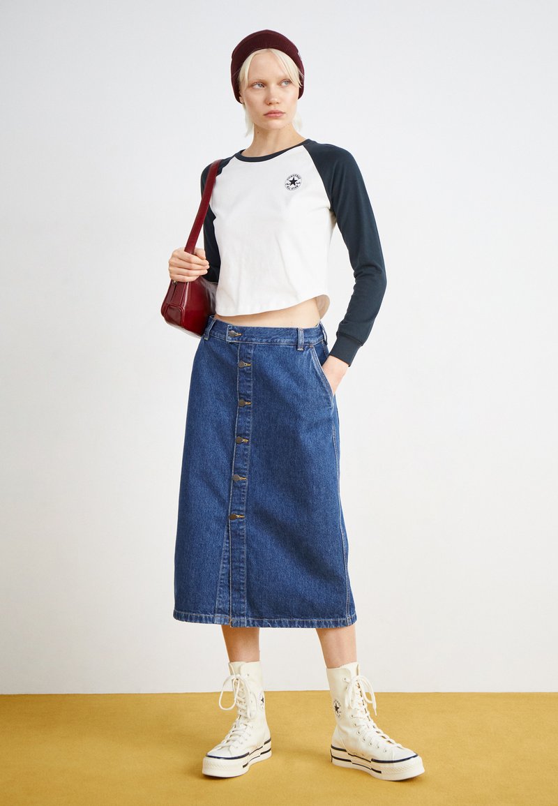 Camicetta bianca e blu a maniche lunghe tagliata, gonna midi in denim blu con bottoni e sneakers alte bianche, abbinate a un berretto bordeaux.