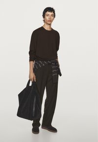 Dunkelbraunes Langarmshirt, grau kariertes Flanellhemd um die Taille gebunden, dunkle Hose, schwarze Wildleder-Sneaker und eine große schwarze Tragetasche.