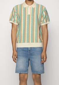 Flerfärgad kortärmad polo med vågiga ränder i pastellnyanser av blått, rosa och kräm, i kombination med blå denimshorts.