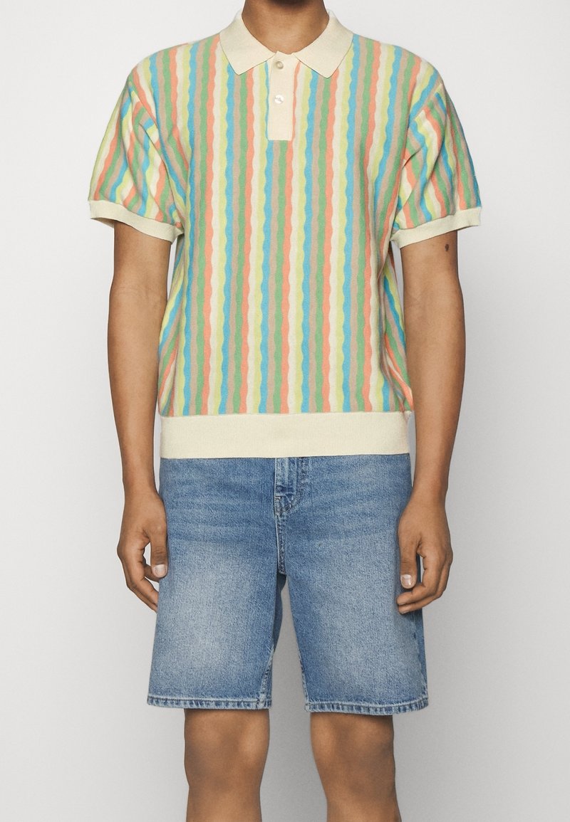 Flerfärgad kortärmad polo med vågiga ränder i pastellnyanser av blått, rosa och kräm, i kombination med blå denimshorts.