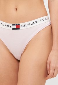 Vaaleanpunainen puuvillabikini, jossa on valkoinen vyötärö, johon on brodeerattu "TOMMY HILFIGER" ja logoaksentteja. Pehmeä rakenne ja yksinkertainen muoto.