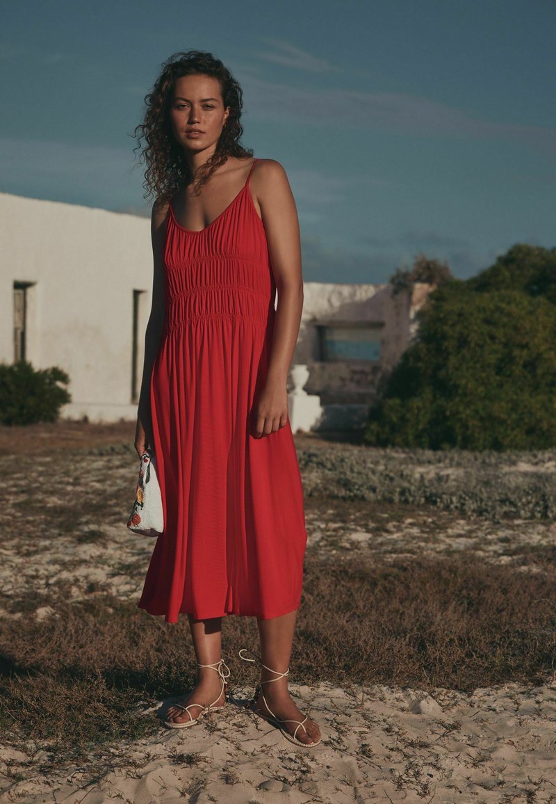 Femme aux cheveux bouclés portant une robe midi rouge sans manches et des sandales blanches se tient dehors à côté d'un bâtiment blanc à la lumière naturelle.