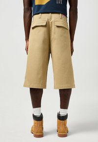 Beige cargoshorts med en løs pasform, der har baglommer og en kant, der når til knæet, parret med brune støvler og hvide sokker.