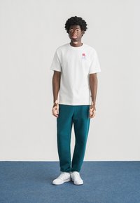 Λευκό βαμβακερό μπλουζάκι με μικρό κόκκινο και μπλε λογότυπο, συνδυασμένο με σκούρο πράσινο φόρμας παντελόνι και λευκά αθλητικά παπούτσια, στέκεται σε μπλε χαλί.