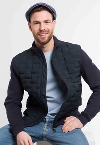 Black padded vest with a zip closure, paired with a navy blue long-sleeve shirt. Casual denim jeans. Model wears a flat cap.