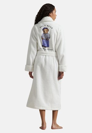 Kvinde iført en hvid fleecebadekåbe med bælte, med et "Polo Bear"-motiv og tekst på bagsiden, stående barfodet på en ensfarvet baggrund.