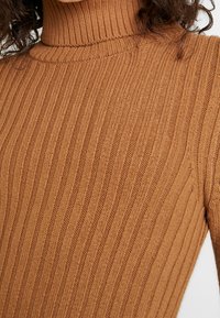 Pull col roulé marron à côtes, mettant en avant une texture verticale et une coupe ajustée, soulignant le matériau tricoté doux et le col haut.