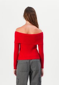 Pull en tricot rouge à épaules dénudées avec une texture côtelée et des manches longues, associé à un pantalon cargo gris avec de grandes poches.