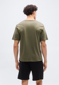 Άνδρας με σγουρά μαλλιά που φοράει ελιάς πράσινη κοντομάνικη μπλούζα  και μαύρες βερμούδες, εμφανιζόμενος από πίσω σε λευκό φόντο.