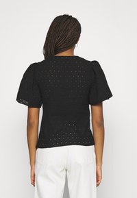 Černý top s krátkými nabíranými rukávy, s výřezovým designem. Materiál je texturovaný a zadní strana má zaoblený výstřih. V kombinaci s bílými kalhotami.