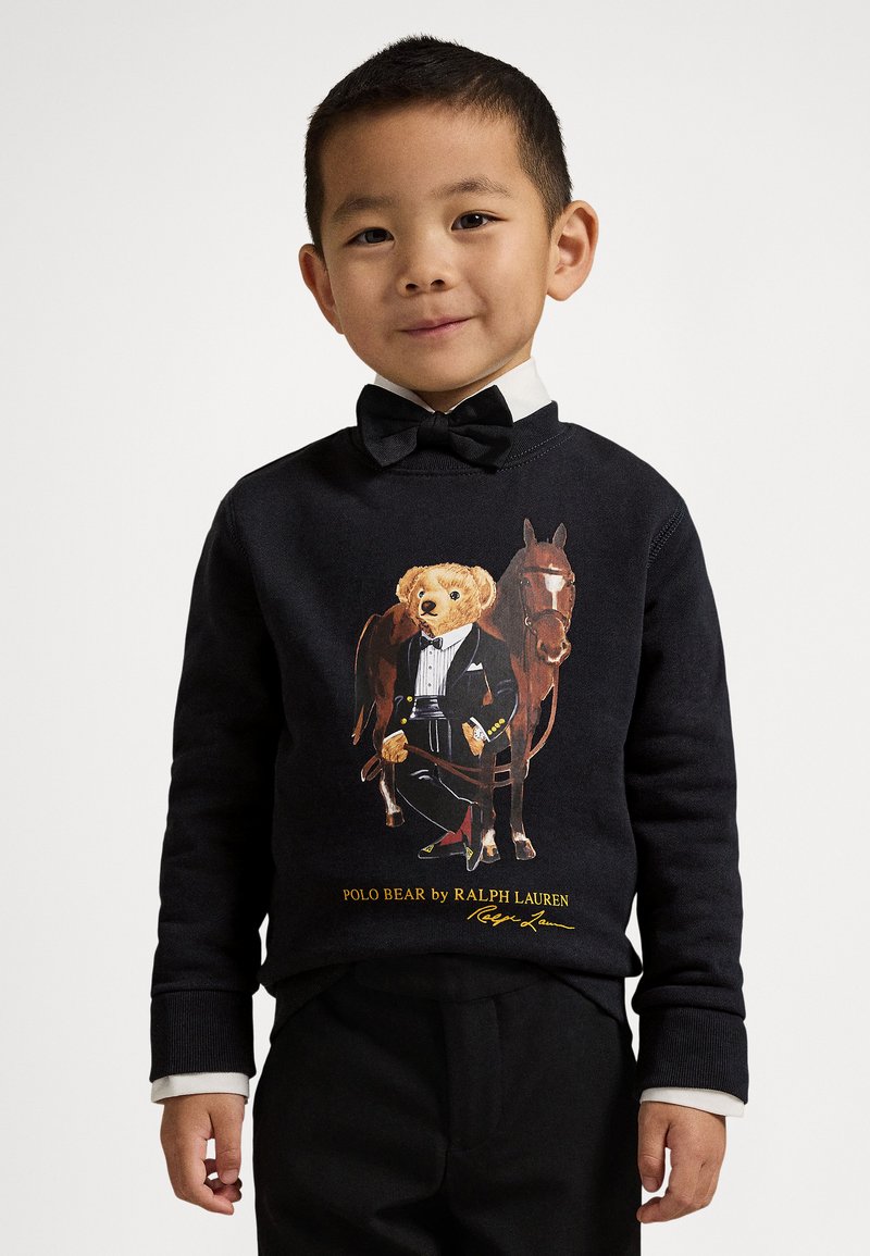 Svart genser med en grafikk av en bjørn i dress ved siden av en hest. Bjørnen holder en stokk, med gullaksenter og bokstaver.