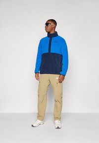 Blauer und marineblauer Fleecepullover mit hohem Kragen, Knopfleiste und Logo. Kombiniert mit khakifarbenen Hosen und weißen Turnschuhen.