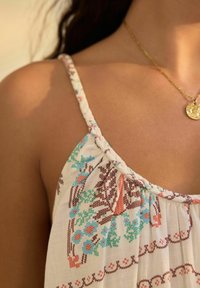 Witte stoffen top met kleurrijke bloemborduursels in teal, oranje en bruin, met dunne gevlochten bandjes en een geplooid ontwerp.