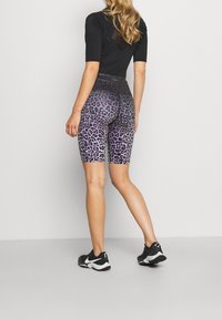 Kobieta w czarnej bluzce z krótkim rękawem i wysokich fioletowych szortach kolarskich w panterkę, z czarnymi i białymi butami sportowymi, stojąca na szarym podłożu.