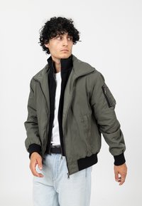 Veste bomber vert olive avec une fermeture éclair frontale, des poignets côtelés et plusieurs poches. Tissu en nylon lisse, design légèrement matelassé, superposé sur une chemise noire.