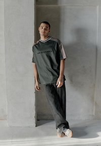 Junger Mann lehnt an einer strukturierten Wand, trägt ein grünes und beiges "Local League"-Shirt, schwarze Jeans und graue Turnschuhe in einem minimalistischen Ambiente.