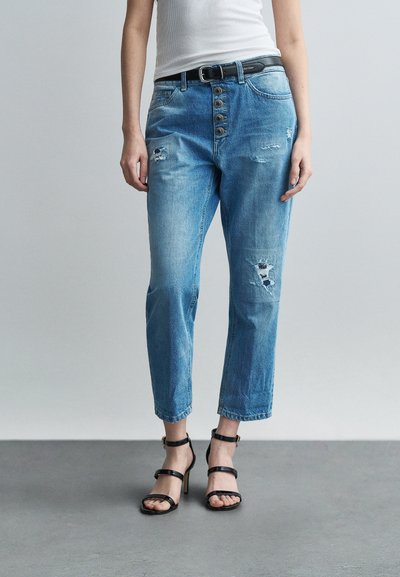 Femme portant un jean taille haute délavé bleu clair avec braguette à boutons et des sandales à talons hauts noires à lanières, debout sur un sol gris.
