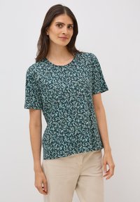 Camisa de manga corta en color teal con un patrón de hojas, cuello redondo, tejido suave y corte relajado, combinada con pantalones beige claros.