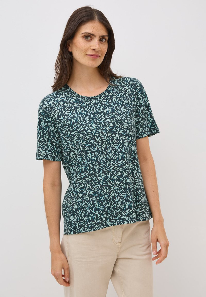 Camisa de manga corta en color teal con un patrón de hojas, cuello redondo, tejido suave y corte relajado, combinada con pantalones beige claros.