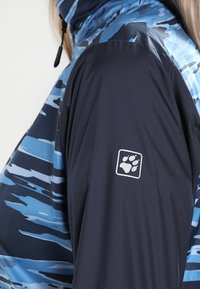 Person i en blå kamouflagemönstrad jacka med svart ärm som har en vit tassavtryck-logotyp på överarmen.