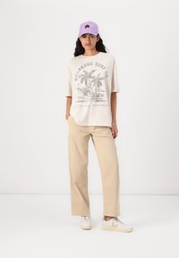 T-shirt en coton beige clair avec un motif de palmier graphique, associé à un pantalon en velours côtelé beige clair et des baskets blanches. Casquette lavande sur la tête.