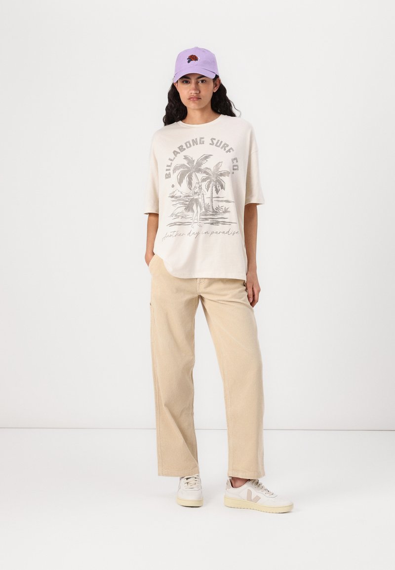 T-shirt en coton beige clair avec un motif de palmier graphique, associé à un pantalon en velours côtelé beige clair et des baskets blanches. Casquette lavande sur la tête.