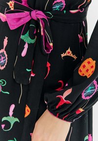 Czarna sukienka z materiału z kolorowymi wzorami, w tym koronami i kształtami. Zawiera różowy pasek do wiązania i elastyczne mankiety na rękawach.