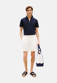 Polo zippé marine, shorts blancs ajustés, sandales foncées, tenant un sac bleu et blanc avec une finition en cuir. Textures lisses, design décontracté.