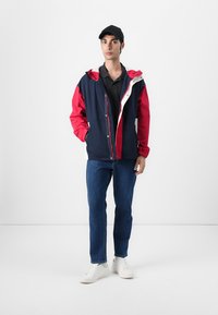 Farveblokeret jakke i marineblå og rød, med en hvid lynlås. Båret over en sort polo, parret med blå jeans og hvide sneakers.