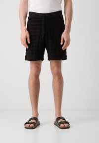 Zwarte shorts van handdoekstof met horizontale strepen, elastische tailleband en trekkoord. In combinatie met zwarte sandalen met een brede band.
