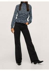 Blouse à manches longues bleu à motif floral avec col montant, associée à un jean noir évasé. La blouse présente une texture froncée à la taille et aux poignets.