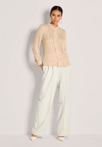 Beige Bluse mit langen Ärmeln und plissiertem Vorderteil, mit Knöpfen und Kragen, kombiniert mit lockeren, hellen Hosen und spitzen Ballerinas.