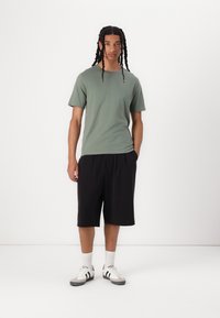 Un tricou cu mâneci scurte de culoare verde măslin asortat cu pantaloni scurți negri până la genunchi, șosete albe și adidași gri cu accente negre, stând pe un fundal alb.