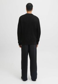 Man die met zijn rug naar de camera staat, gekleed in een zwart gemêleerd sweater en losse zwarte broek, tegen een eenvoudige lichte achtergrond.