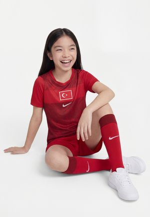 Fille souriante assise par terre portant un uniforme de football turc rouge, des chaussettes rouges et des baskets blanches Nike sur fond blanc.