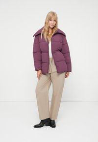 Paarse pufferjack met een brede kraag, pluche textuur, een rits aan de voorkant en een elastische zoom, gecombineerd met beige broeken en zwarte enkellaarsjes met hak.