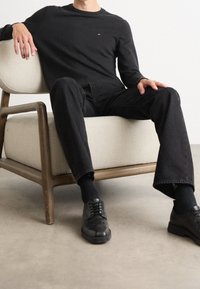 Camicetta nera a maniche lunghe con piccolo logo, jeans neri, calzini neri e scarpe. Seduto su una sedia di tessuto chiaro con telaio in legno.