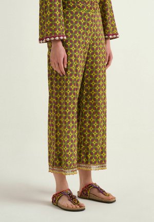 Persona che indossa pantaloni a gamba larga e top abbinato con motivo giallo e viola, in piedi a piedi nudi con sandali decorati su sfondo chiaro.