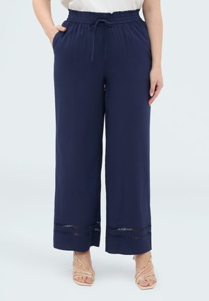 Kobieta nosząca granatowe szerokie spodnie z elastycznym pasem i koronkową lamówką na nogawkach, zestawione z beżowymi sandałami na obcasie z paskami.