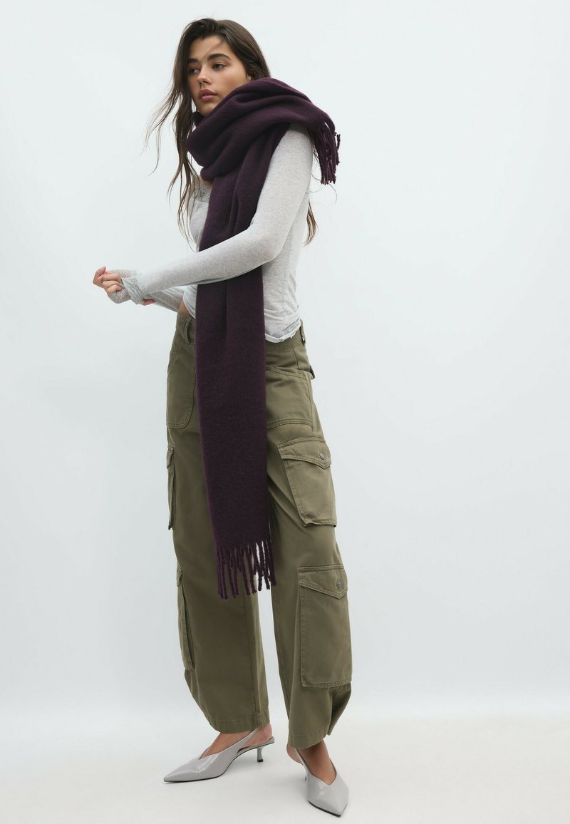 Femme portant un haut gris clair à manches longues, une grande écharpe violet foncé, un pantalon cargo vert olive et des chaussures grises à petits talons, debout devant un fond blanc.