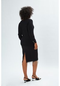 Schwarzes Kleid mit langen Ärmeln, seitlichem Schlitz am Saum, verziert mit Fransen. Getragen mit schwarzen Slingback-Absätzen, die dekorative Akzente aufweisen.