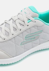 Sneakers gris clair avec une tige texturée et matelassée. Présentent des accents et des lacets turquoise, et une semelle en caoutchouc blanche. Logo affiché sur le côté.