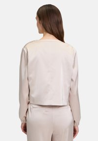 Kurze beigefarbene Satinjacke mit langen Ärmeln, rundem Halsausschnitt und tailliertem Design. Glatte Textur und minimalistischer Stil. Keine sichtbaren Verschlüsse.