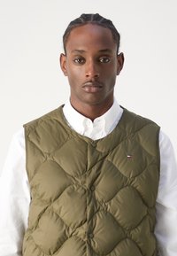 Olivenfarvet quiltet vest med cirkulært mønster, trykknapper og lille logo på brystet, lagdelt over en hvid skjorte med krave.
