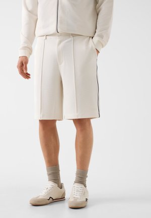 Uomo che indossa pantaloncini color bianco sporco con tasche laterali, abbinati a calzini color taupe e sneaker bianche su uno sfondo semplice.