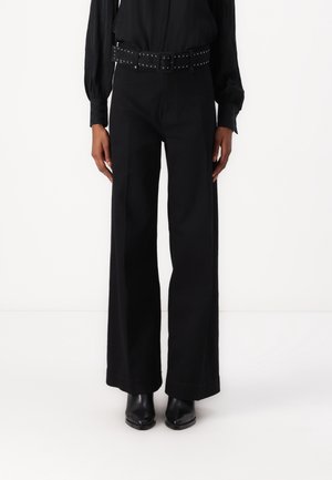 Person trägt hoch taillierte, weit geschnittene schwarze Hose, schwarzen Nietengürtel, schwarze langärmlige Bluse und schwarze spitze Stiefel.