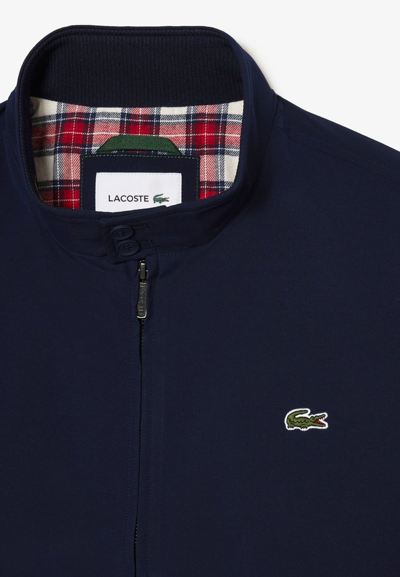 Veste bleu marine avec un col haut, fermeture éclair à l'avant et deux boutons. L'intérieur présente une doublure à carreaux rouge et blanc. Logo Lacoste brodé.