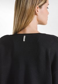 Maglione nero con scollo ampio, caratterizzato da una finitura a maglia testurizzata e un piccolo etichetta bianca sul retro. Design semplice e moderno.