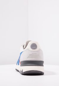 Sneaker con tomaia in suede grigia e rete bianca, dettagli blu e rossi, suola in gomma nera e un particolare tondo grigio sul tallone.